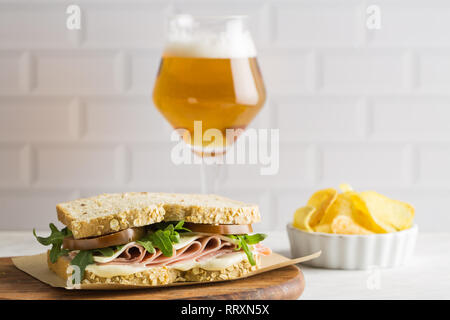 Lecker Sandwich mit Schinken, Käse, Salat und Tomaten mit einem Glas Bier und Chips auf hellen Hintergrund Stockfoto