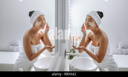 Weibliche in Handtuch Anwendung Creme auf ihrem Gesicht. Asiatische Frau im Bad in den Spiegel und Anwendung der Creme auf ihrem Gesicht. Stockfoto