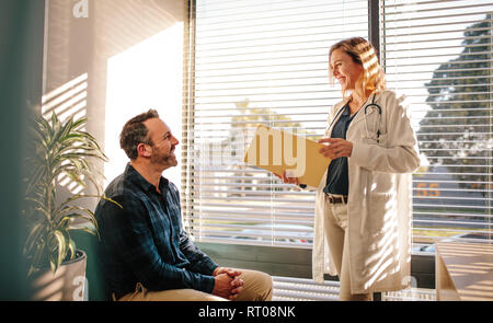 Lächelnd weibliche Arzt Holding einen medizinischen Bericht Datei und im Gespräch mit männlicher Patient sitzt auf einem Sofa am Krankenhaus Rezeption. Arzt teilen gute medizinische t Stockfoto