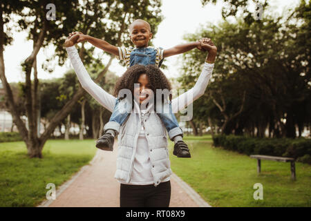 Glückliche Mutter und Sohn ein piggyback Ride auf ihren Schultern im Park. Afrikanische Frau, die seine Jungen auf ihre Schultern beim Gehen in einen Park. Stockfoto