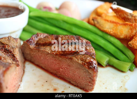 Geschlossen bis medium Gegrillte Filet Steak in der Hälfte mit verschwommenen gedünstetem Gemüse und Bratkartoffeln serviert auf weiße Platte Stockfoto