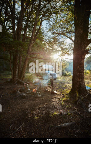 Chile, Santa Magda, Rio Maniguales, Junge am Lagerfeuer sitzen im Wald mit Wohnmobil im Hintergrund Stockfoto