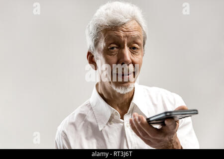 Indoor Portrait von attraktiven älteren Mann auf grauem Hintergrund isoliert, leer, Smartphone, Sprachsteuerung, Gefühl, glücklich und überrascht. Menschliche Gefühle, Mimik Konzept. Stockfoto
