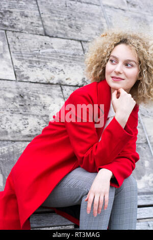 Portrait von lächelnden blonde Frau mit korkenzieherlocken das Tragen der roten Mantel beobachten etwas Stockfoto