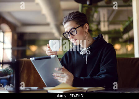 Smart qualifizierten Studenten mit kurzen Haaren in modischen Brillen trinken Kaffee und Studieren mit modernen Tablet in der Cafeteria Stockfoto