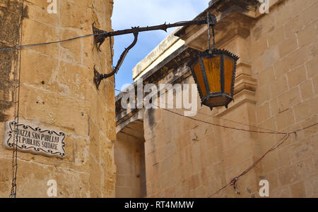 Panoramablick auf einem alten, antiken, mittelalterlichen und historischen Laterne hängend auf einer sand Steinmauer. Im Hintergrund das Schloss von Mdina, Malta. Europa Stockfoto