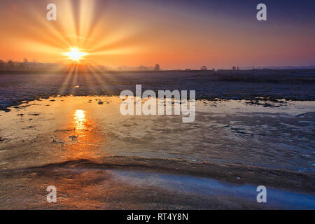 Sonnenaufgang über gefrorene Felder und die Reflexion der Sonne in der Eisschicht auf dem Feld. Polska Ende Februar. horizontale Ansicht. Stockfoto