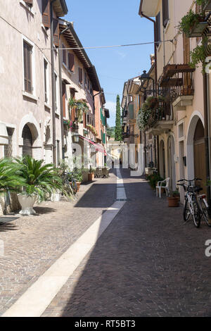 BARDOLINO, Venetien, Italien - 7. AUGUST 2017: Schmale Straße mit Restaurants und Geschäften mit bunten Fassaden der Wohnhäuser und blauer Himmel. Stockfoto