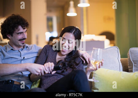 Verspieltes Paar kämpfen um die Fernbedienung, Fernsehen und das Trinken von Wein auf dem Sofa Stockfoto