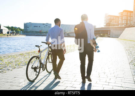 Zwei Geschäftsleute gehen mit Fahrrad und Skateboard am Flussufer Stockfoto