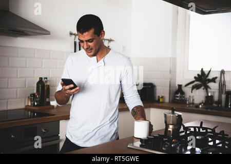 Lächelnden jungen Mann mit Tasse Kaffee mit Handy in der Küche zu Hause Stockfoto