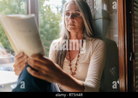 Portrait von Frau auf Fensterbank, zu Hause zu sitzen, ein Buch lesen Stockfoto