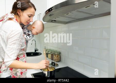 Junge Mutter mit Baby auf ihren Händen kocht das Essen in einem Topf auf dem Herd Stockfoto