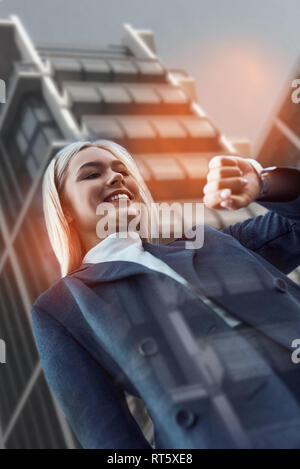 Welche Zeit ist es? Fröhliche Business woman in der Klage auf ihre Uhr zu schauen und zu lächeln, während sich gegen moderne Bürogebäude. Zeit-management. Zeit Konzept. Geschäftsreise Stockfoto