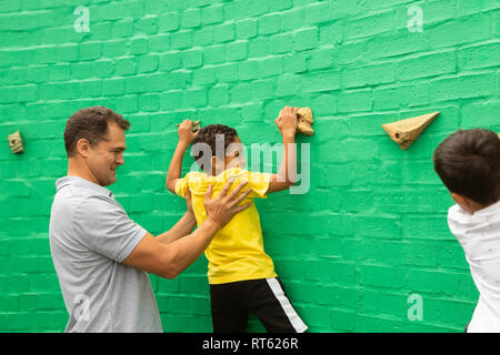 Männliche Lehrer Unterstützung ein schuljunge künstlichen Mauer zu klettern Stockfoto