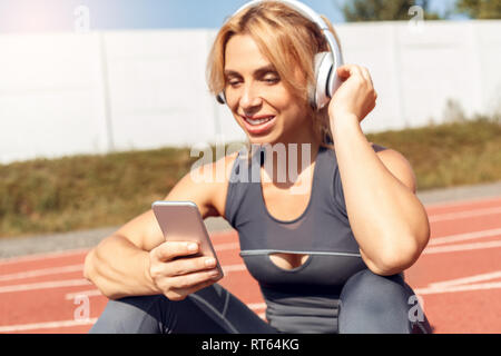 Sportlicher Lifestyle. Junge Frau in Kopfhörer im Stadion sitzen am Anschluss chosing Song auf dem Smartphone Musik glücklich close-up Stockfoto