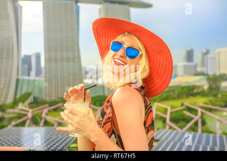 Reisen Urlaub Ferien in Singapur, Südostasien. Lifestyle kaukasische Frau mit breiten Hut trinken Aperitif auf der Dachterrasse. Luftbild oder Stadtbild Stockfoto