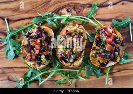 Frisches Bruschetta mit einer Vielzahl von Oliven und würfelt Tomaten auf kleinen getoasteten Brotscheiben mit Balsamico Reduzierung auf einer hölzernen Fach beträufelt gemacht Stockfoto