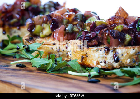 Frisches Bruschetta mit einer Vielzahl von Oliven und würfelt Tomaten auf kleinen getoasteten Brotscheiben mit Balsamico Reduzierung auf einer hölzernen Fach beträufelt gemacht Stockfoto