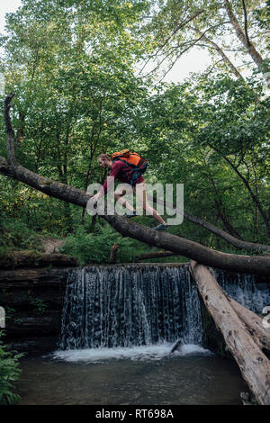 Junge Wanderer mit Rucksack Kreuzung Wasser auf Baumstamm im Wald Stockfoto