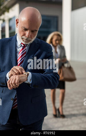 Senior Geschäftsmann Kontrolle der Zeit mit Geschäftsfrau im Hintergrund Stockfoto