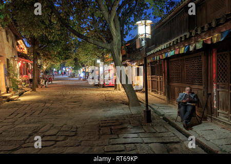 Shaxi, China - 21. Februar 2019: Chinesischer Mann reasting auf einen Stuhl in der zentrale Platz von shaxi Altstadt in der Dämmerung Stockfoto