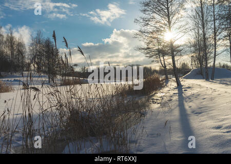 Gefrorenen Schnee bedeckten Fluss in einer Winterlandschaft mit Sunburst durch die Wolken zurück Beleuchtung Schilf auf der Bank Stockfoto