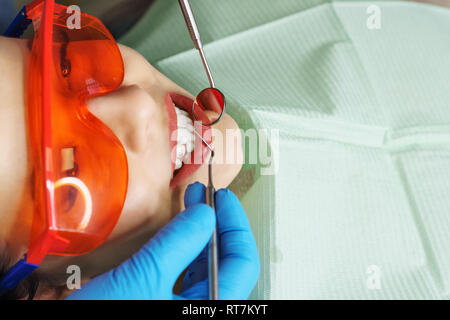 Vorbeugende Prüfung an Zahnarzt. Junge Mädchen in der Schutzbrille ist ein Lächeln auf den Lippen. Weiße Zähne. Der Zahnarzt die Hände in Handschuhe mit medizinischen instrum Stockfoto