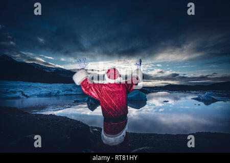 Island, Ansicht von hinten von einem Mann verkleidet als Weihnachtsmann stehend auf einem Gletscher seine Arme anheben Stockfoto