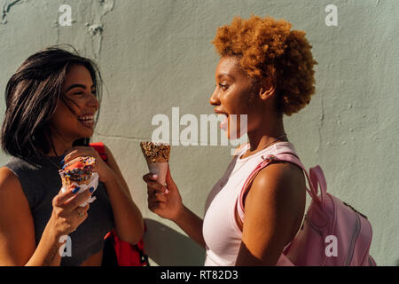 Zwei glückliche weibliche Freunde mit Eistüten Gespräch an einer Wand Stockfoto