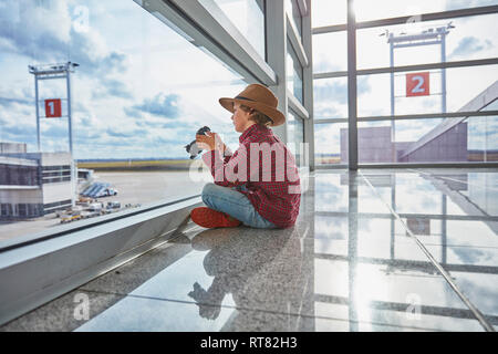 Junge sitzt hinter Fensterglas am Flughafen halten einer Kamera Stockfoto