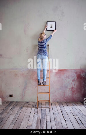 Rückansicht der Frau auf Leiter aufhängen Bilderrahmen in einem unrenovierten Zimmer eines Loft Stockfoto