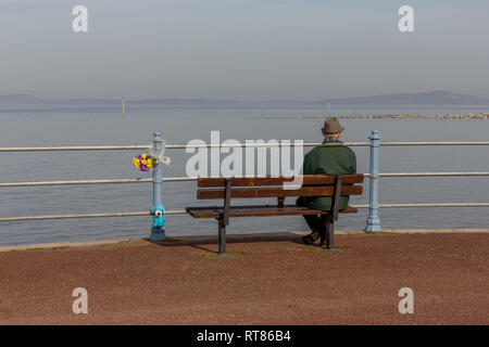 Ein einsamer Mann saß auf einer Promenade Sitzbank in Morecambe Bay suchen neben einem floralen Tribut Stockfoto