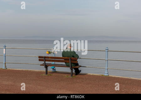 Ein einsamer Mann saß auf einer Promenade Sitzbank in Morecambe Bay suchen neben einem floralen Tribut Stockfoto