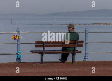 Ein einsamer Mann saß auf einer Promenade Sitzbank in Morecambe Bay suchen neben einem floralen Tribut Stockfoto