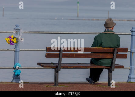 Ein einsamer Mann saß auf einer Promenade Sitzbank in Morecambe Bay suchen neben einem floralen Tribut Stockfoto