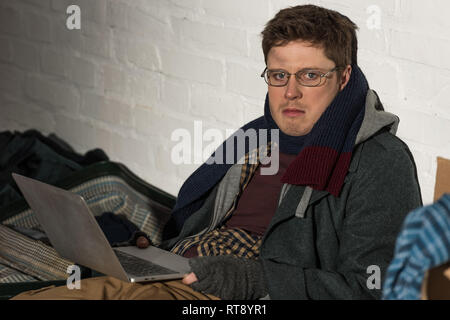 Obdachloser in Gläsern mit Laptop, während durch weiße Mauer sitzen Stockfoto