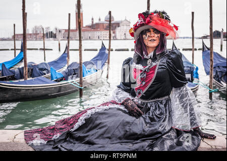 Schöne maskierte Frau während des Karnevals in Venedig (Italien) in der Nähe der Lagune mit vielen Gondeln an der Rückseite Stockfoto