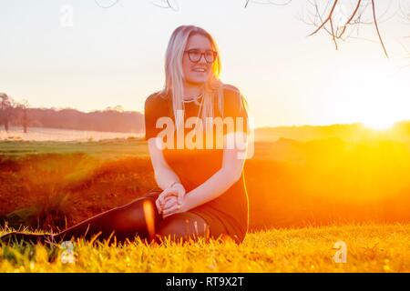 Schöne Frau sitzt im Gras vor Sonnenuntergang aus der Blick in die Ferne lächelte zufrieden. Stockfoto