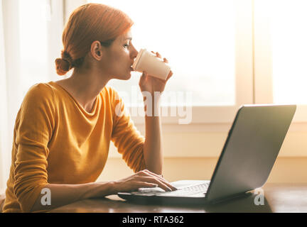 Junge Frau trinkt Kaffee zum Mitnehmen während der Arbeit am Laptop Stockfoto
