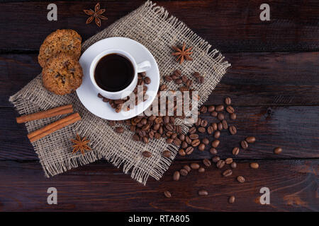 Weiße Tasse Kaffee mit Kaffee Körner in der Nähe und Keksen auf Leinen Stoff auf der dunklen vintage Hintergrund. Stockfoto