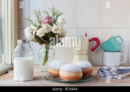 Drei frische Donuts auf eine Glasplatte mit Kaffeetassen, moka Topf, Blumen und Milch Flasche im Hintergrund, durch das Fenster Stockfoto