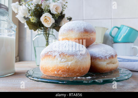 Drei frische Donuts auf eine Glasplatte mit Kaffeetassen, die Blumen und die Milchflasche im Hintergrund Stockfoto