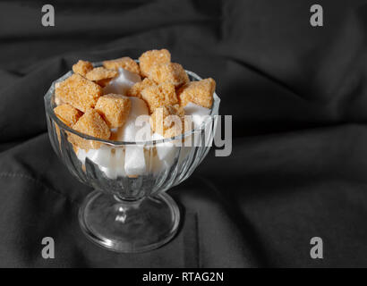 Würfel weißen und braunen Zucker in einem Glas Vase auf dem Hintergrund der dunklen Tuch. Vorhänge. Selektive konzentrieren. Rechteckiges Bild. Stockfoto