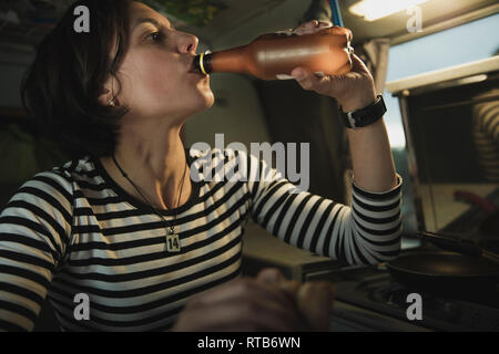 Junge brünette Dame Trinkwasser aus der Flasche in der Nähe von Herd in Mobile home Stockfoto