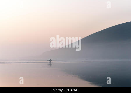 Ein einsamer Surfer zu Fuß am Strand entlang auf einem nebligen Winter am Nachmittag. Saunton, Devon, Großbritannien. Stockfoto