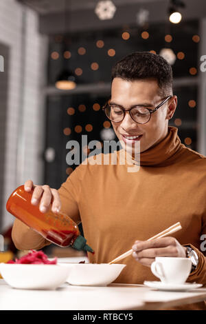 Junger Mann hinzufügen Ketchup zu essen, während Sie die Stäbchen mit der anderen Hand. Stockfoto