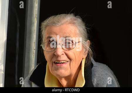 Porträt der fröhliche, gesunde alte Dame mit Brille schauen durch Fenster Stockfoto