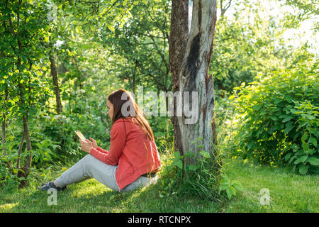 Mädchen in einem sportlichen rosa Jacke und graue Hose sitzt auf einem grünen Frühling Rasen in der Nähe von einem Baum und liest auf dem Telefon Stockfoto