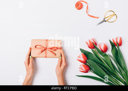 Die Hände oben Frau mit handgefertigten verpackte Geschenk neben Bouquet von roten Tulpen, Satinband und goldenen Schere, Flachbild festliche Komposition legen. Stockfoto